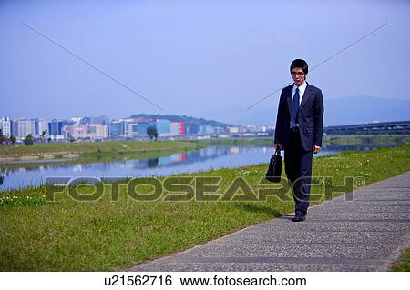 Stock Photograph - Young businessman holding briefcase walking on footpath. Fotosearch