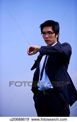 Young businessman looking at watch against blue sky View Large Photo Image Stock Photo - Young businessman looking at watch against blue sky. Fotosearch