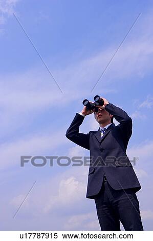 Stock Photography - Young businessman looking through binoculars, standing against sky. Fotosearch