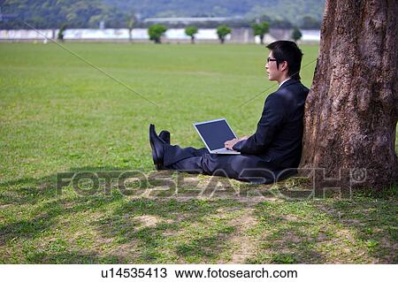 Stock Image - Young businessman with laptop leaning against tree. Fotosearch