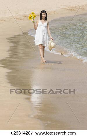 Stock Photo - "Young woman holding sunflower and sunhat, walking on beach". Fotosearch