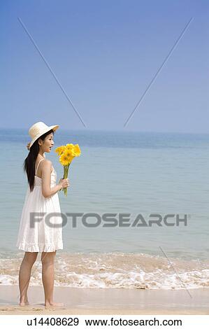 Stock Photo - Young woman holding sunflowers and standing on the beach. Fotosearch