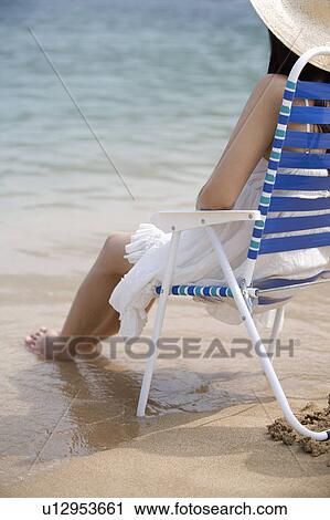 Stock Image - Young woman sitting on beach chair with feet in water. Fotosearch