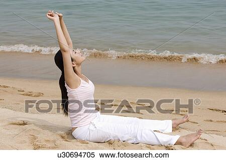 Stock Photography - Young woman sitting on beach with arms outstretched. Fotosearch
