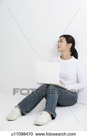 Stock Image - "Young woman sitting on the floor, using laptop and looking away". Fotosearch