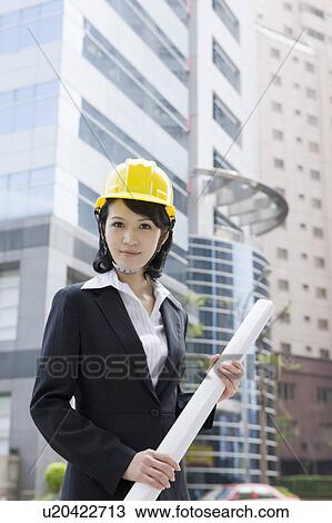 Stock Image - Businesswoman holding blueprint and looking at the camera with helmet. Fotosearch