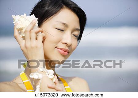 Stock Image - Young woman holding seashell to ear. Fotosearch