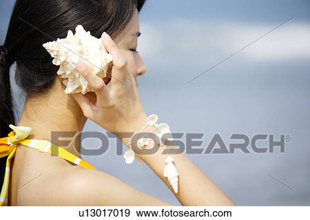 Stock Photo - Young woman holding seashell to ear, side view. Fotosearch