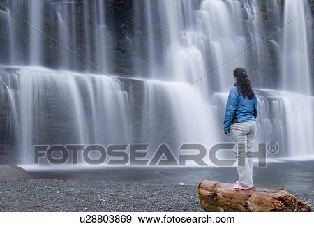 Stock Photo - Woman (20-25) standing on a beach log in front of a waterfall. Fotosearch