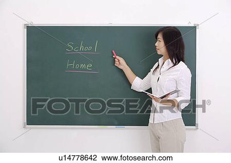 Stock Image - Teacher pointing at the blackboard and holding a textbook. Fotosearch