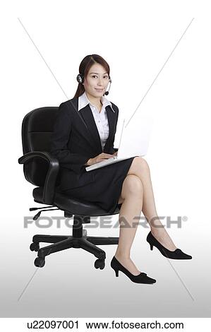 Stock Image - Young business woman sitting on the office chair and wearing the headphones. Fotosearch