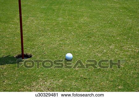 Stock Image - Golf ball beside hole containing flag pole. Fotosearch