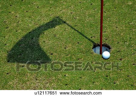 Stock Image - Golf ball perched at edge of hole containing flag pole. Fotosearch