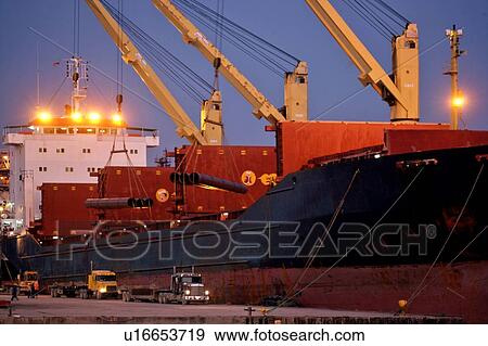 Stock Photo - Offloading Pipe from Cargo Ship to Truck at Dusk. Fotosearch