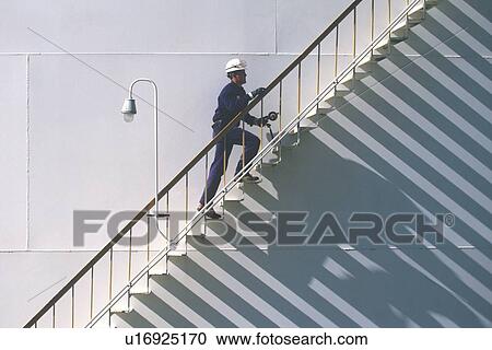 Stock Image - Oil Refinery Worker Climbing Storage Tank Stairway. Fotosearch
