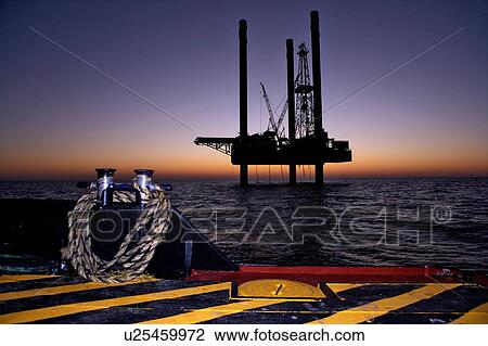 Stock Image - Silhouete View at Sunset of Offshore Oil Drilling Rig from Supply Boat. Fotosearch