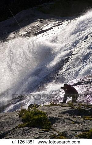 Stock Image - A photographer at a snow melt waterfall in Nordfjord, an arm of the larger Melfjord - situated just above the Arctic Circle - in northern Norway, Barents Sea.. Fotosearch