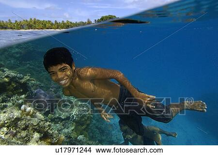 Picture - Over under image of Marshallese boy smiling underwater next to coral reef and coconut trees lining the shore of Majikin Island, Namu atoll, Marshall Islands (N. Pacific). Fotosearch