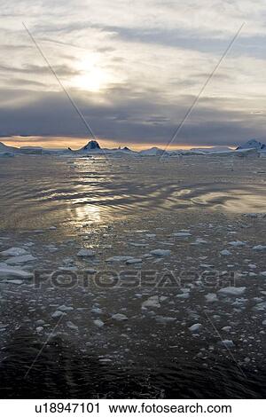 Stock Image - Sunset on snow-capped mountains and icebergs surrounded in brash ice in Crystal Sound near the Antarctic Circle on the west side of the Antarctic Peninsula.. Fotosearch