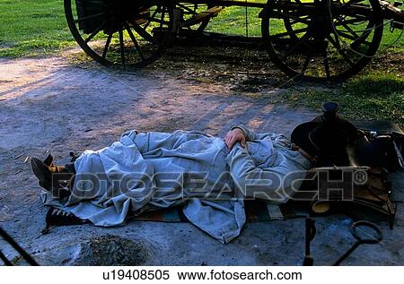 Stock Photography - man sleeping udner blankets by a covered wagon. Fotosearch