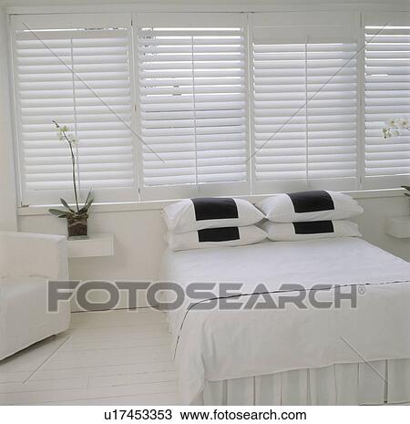 Stock Image - All white bedroom with colonial style shutters, blackand white pillows and painted floor.. Fotosearch