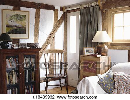 Stock Image - Antique bookcase and wooden chair in cottage livingroom with exposed wall beams. Fotosearch