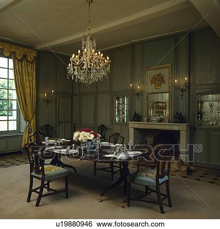 Stock Photograph - Antique crystal chandelier above antique table and chairs in green panelled country dining room with cream carpet. Fotosearch