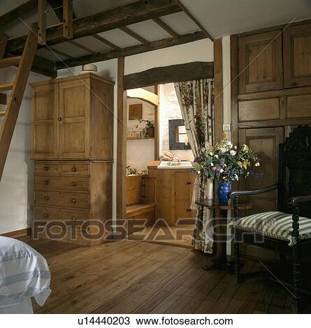 Stock Image - Antique linen press beside doorway with curtain to ensuite bathroom in country bedroom with wooden flooring. Fotosearch