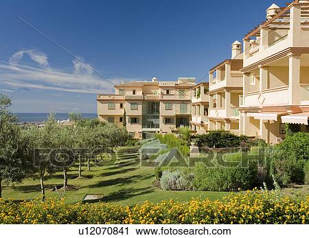 Stock Image - Avenue of small trees in gardens in front of neutral modern apartment building by the ocean in southern Spain. Fotosearch