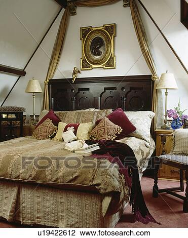 Stock Image - Bedroom with ornate gilt-framed picture above carved bed with patterned beige bedlinen and red velvet throw. Fotosearch