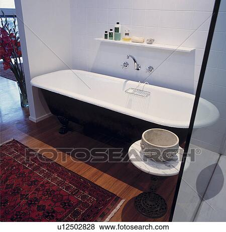 Stock Photo - Black rolltop bath and oriental rug on wooden floor in modern white tiled bathroom. Fotosearch