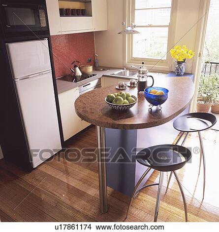 Picture - Black stools at oval breakfast bar with cafetiere in modern kitchen with fridge-freezer and wooden flooring. Fotosearch