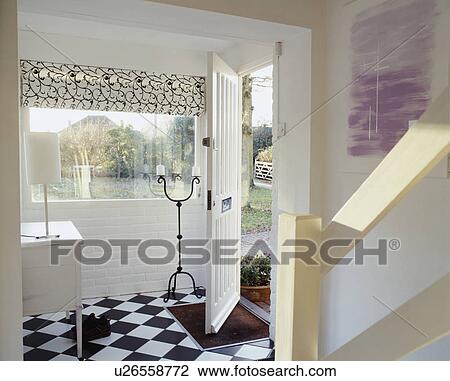 Stock Image - Black+white blind on window beside open door in small modern white hall with black+white chequerboard floor. Fotosearch