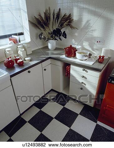 Stock Image - Black+white chequerboard vinyl flooring in small white kitchen with red crockery on granite worktop with white hob. Fotosearch