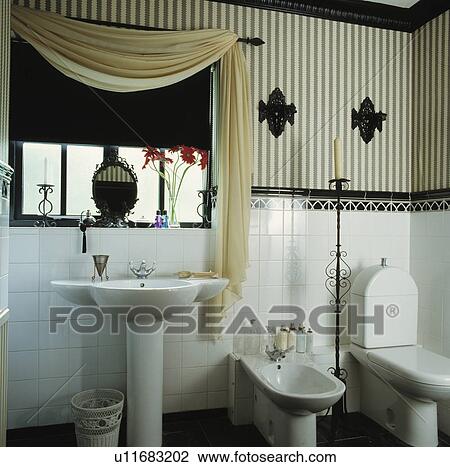 Stock Image - Black+white striped wallpaper above white tiled wall in bathroom with cream drape and black blind on window. Fotosearch