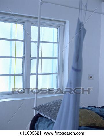 Stock Image - Blue dress hanging from white metal bed in front of window in white bedroom. Fotosearch
