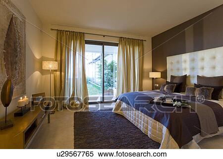 Stock Photography - Blue rug and patterned bedlinen in modern apartment bedroom with sheer neutral curtains at patio doors. Fotosearch