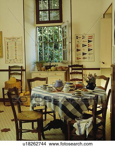 Picture - Blue+white checked cloth on table with rush-seated chairs in cream country kitchen with security bars on open window. Fotosearch