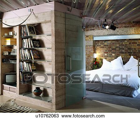 Stock Image - Books and television on wooden unit beside glass door to modern bedroom with brick walls. Fotosearch