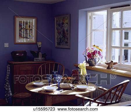 Stock Image - Bowls and blue glasses on circular pine table with Windosr chairs iin front of window in blue cottage dining room. Fotosearch