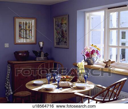 Bowls and blue glasses on circular pine table with Windosr chairs iin front of window in blue cottage dining room View Large Photo Image Stock Photograph - Bowls and blue glasses on circular pine table with Windosr chairs iin front of window in blue cottage dining room. Fotosearch