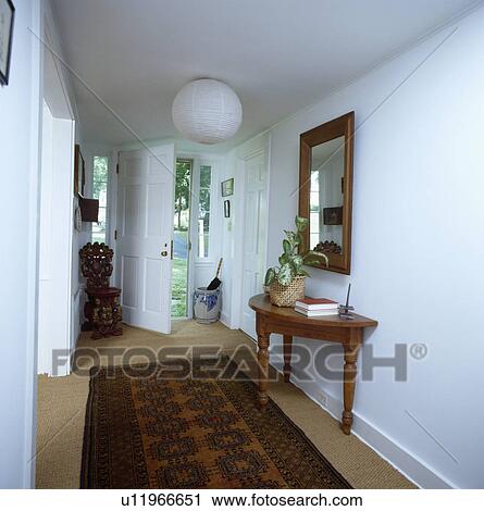 Stock Image - Brown oriental rug on sisal carpet in white country hall with paper globe lampshade and mirror above console table. Fotosearch