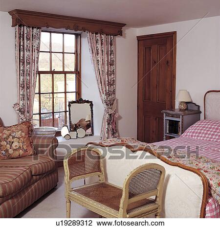 Stock Image - Cane stool below bed with checked duvet and floral quilt in bedroom with floral curtains below wooden pelmet on window. Fotosearch