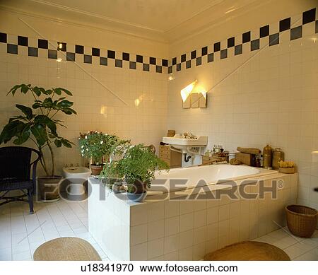 Stock Image - Central bath with white tiled surround in modern white tiled bathroom with black+white tiled chequerboard border . Fotosearch