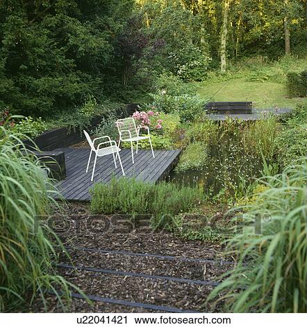 Stock Image - Chairs on decked patio beside rectangular pond in large country garden. Fotosearch