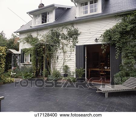 Stock Image - Chaiselongue on slate patio in front of traditional clapboard house in France. Fotosearch
