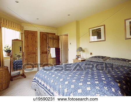 Stock Image - Cheval mirror and blue patterned duvet on bed in pastel yellow traditional bedroom. Fotosearch