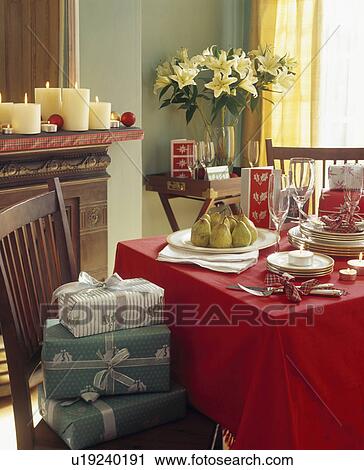 Stock Image - Christmas presents on chair in diningroom with white crockery on red tablecloth. Fotosearch