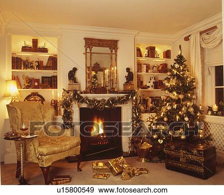Stock Photo - Christmas tree and armchair beside fireplace in gold-themed living room. Fotosearch