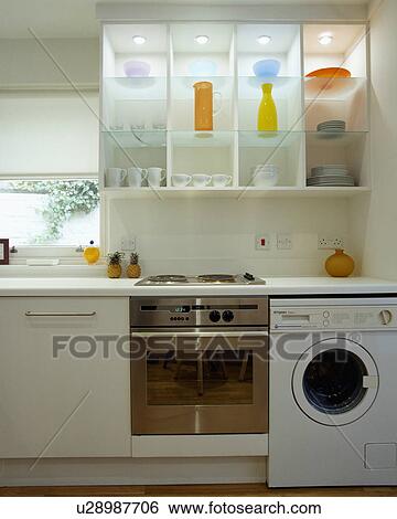Stock Photograph - Chrome oven and white washingmachine below lighted glass shelves with coloured vases in modern white kitchen. Fotosearch
